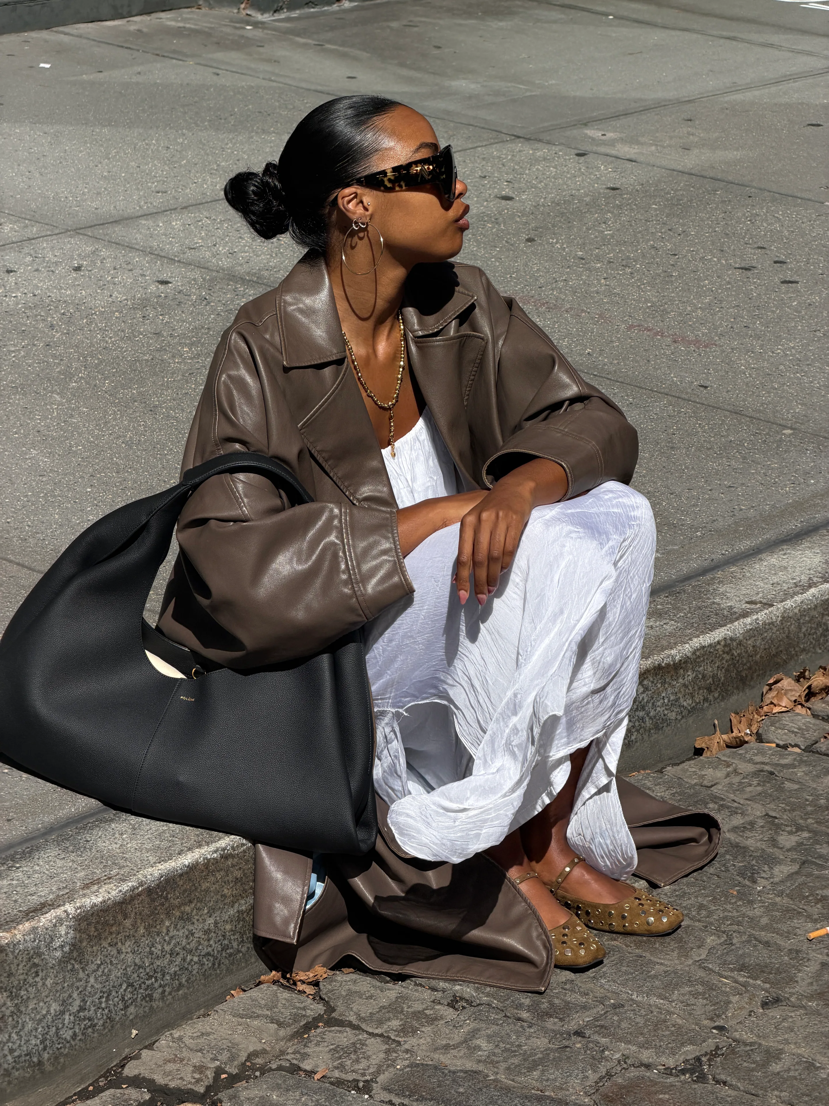 Brown leather trench coat over white dress, gold necklace, studded flats, and black handbag.