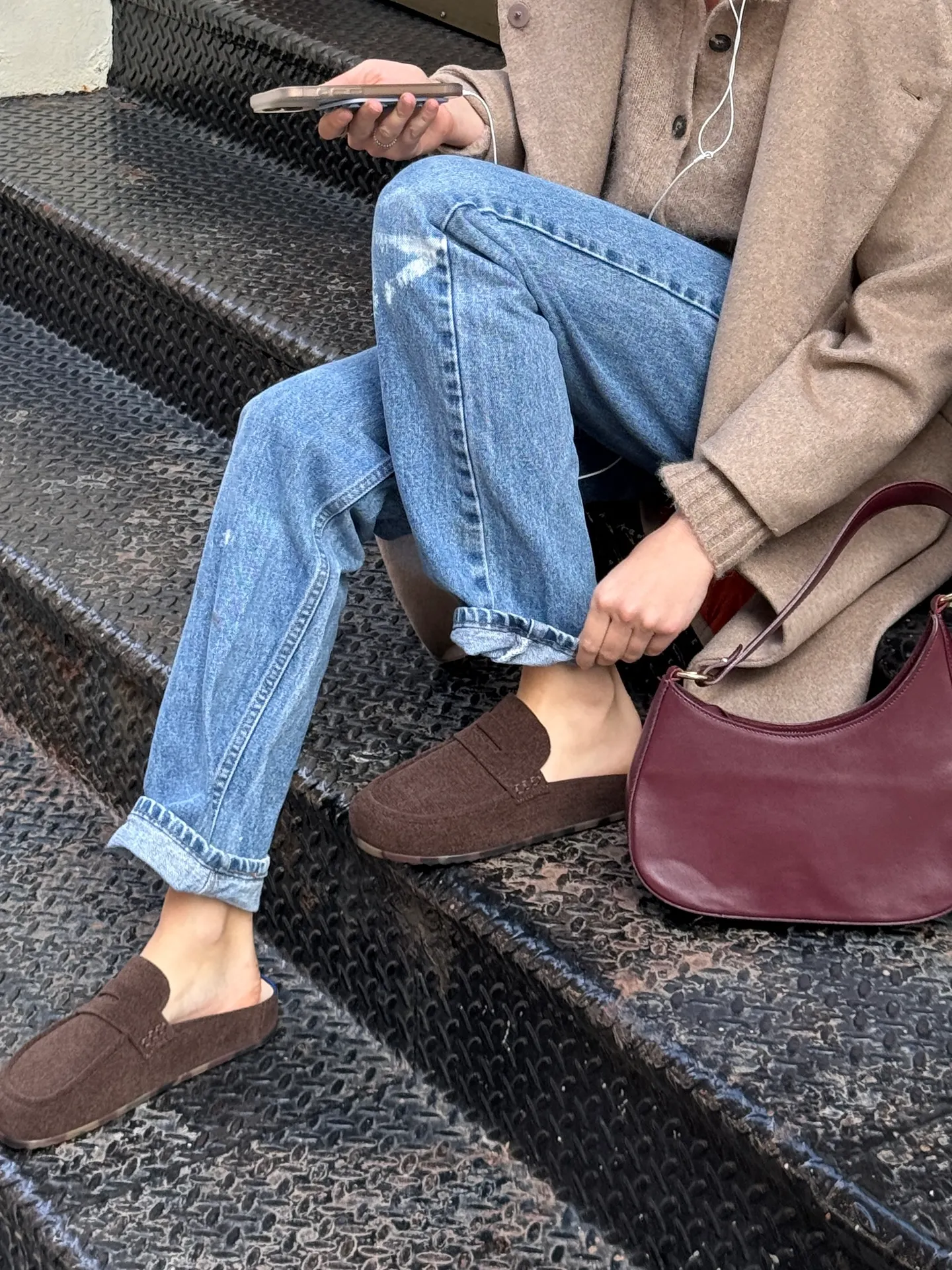 Beige coat, light wash jeans, brown slip-on loafers, and a burgundy shoulder bag on stairs.
